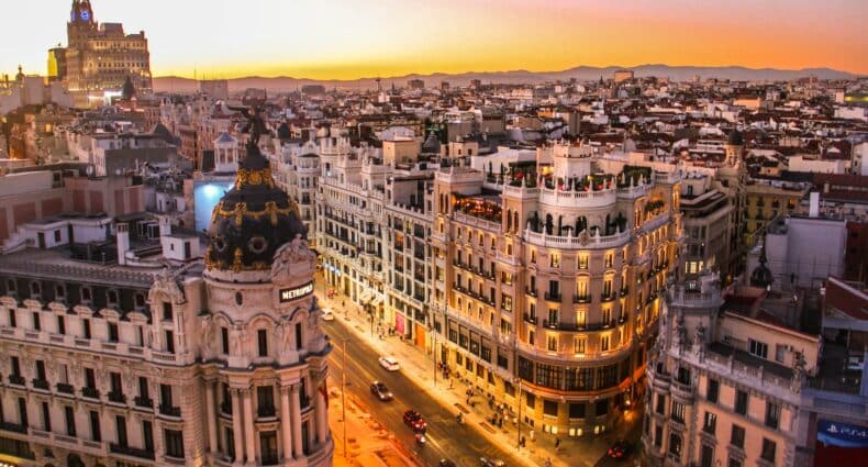 Fotografia aérea da Calle Gran Via em Madri ao pôr do Sol. Prédios luxuosos estão iluminados e ladeiam uma rua onde passam carros. Ao fundo é possível ver outros prédios e construções. - Foto: Florian Wehde via Unsplash