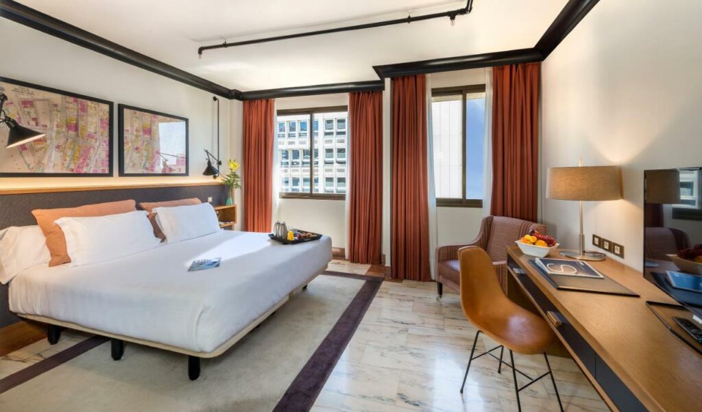 Quarto do H10 Tribeca, uma das recomendações de hotéis baratos em Madri. Uma cama de casal tem mesinhas de cabeceira e luminárias dos dois lados encara uma mesa de trabalho com cadeira, abajur, tigela com frutas e TV. Uma poltrona marrom está perto da parede que tem duas janelas com cortinas. Há uma bandeja de café da manhã aos pés da cama.