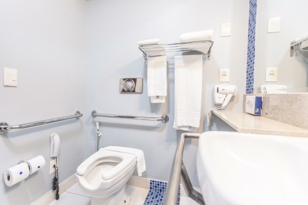 Banheiro do hotel Holiday Inn Cuiaba com duas barras de apoio ao redor do vaso, uma ao redor da pia, toalhas e um telefone na altura do vaso.