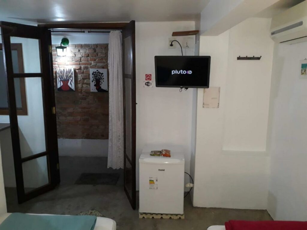 Vista do outro lado de um quarto na Hospedagem Casa de Pedro. Há um frigobar, uma televisão pendurada na parede, ar-condicionado e uma porta dando acesso ao corredor.