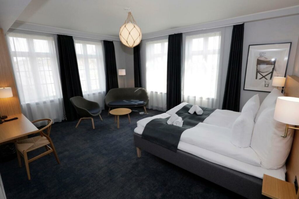 Quarto do Hotel Bethel com duas camas juntas formando uma cama de casal, cadeiras, várias janelas com cortinas e uma mesa. Foto para ilustrar post sobre hotéis em Copenhague.