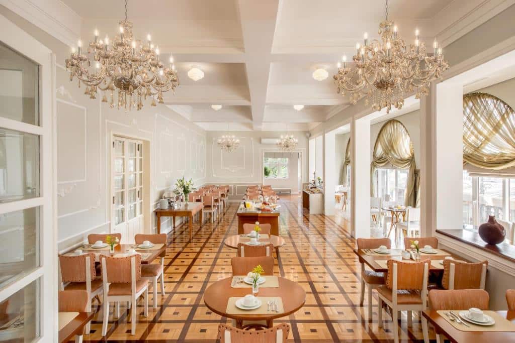 Salão de refeições do Hotel Casacurta, com candelabros no teto alto, e mesinhas diversas, redondas e quadradas, espalhadas pelo espaço. A décor é retrô, e há espaço para caminhar entre as mesas.
