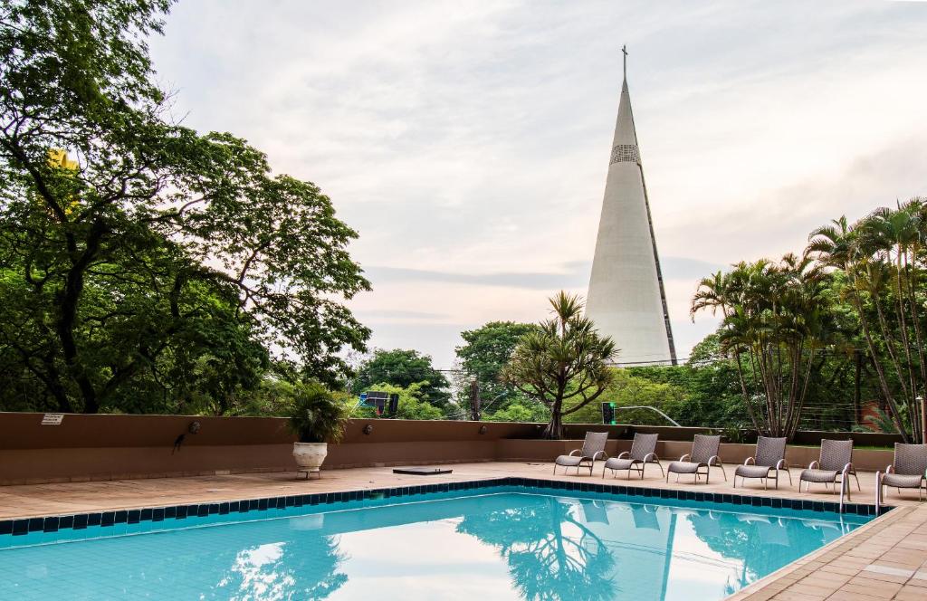 Parte da piscina azul com algumas cadeiras e árvores verdes em volta, ao fundo uma construção triangular durante o dia, ilustrando post Hotéis em Maringá.