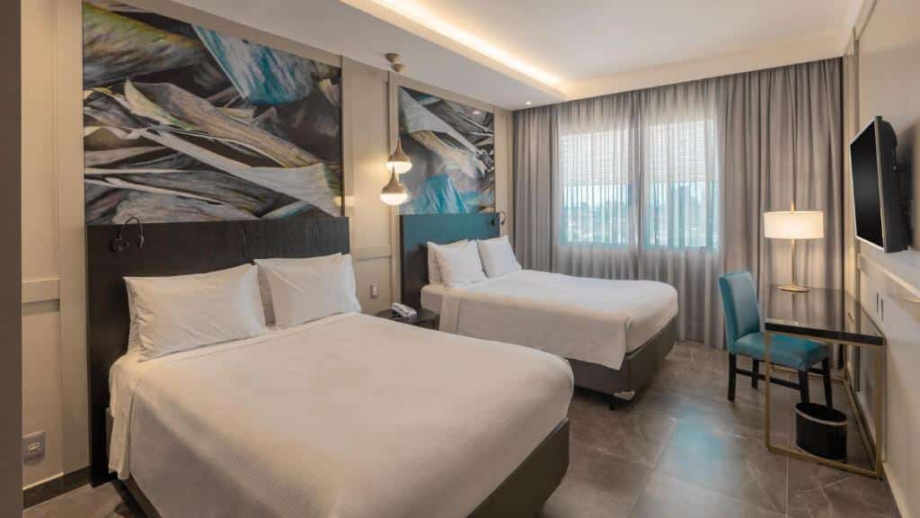 Quarto do hotel Deville Prime Cuiabá com duas camas de casal. Há também uma mesa com cadeira e abajur, luminárias e uma janela com cortina. Foto para ilustrar post sobre hotéis em Cuiabá.