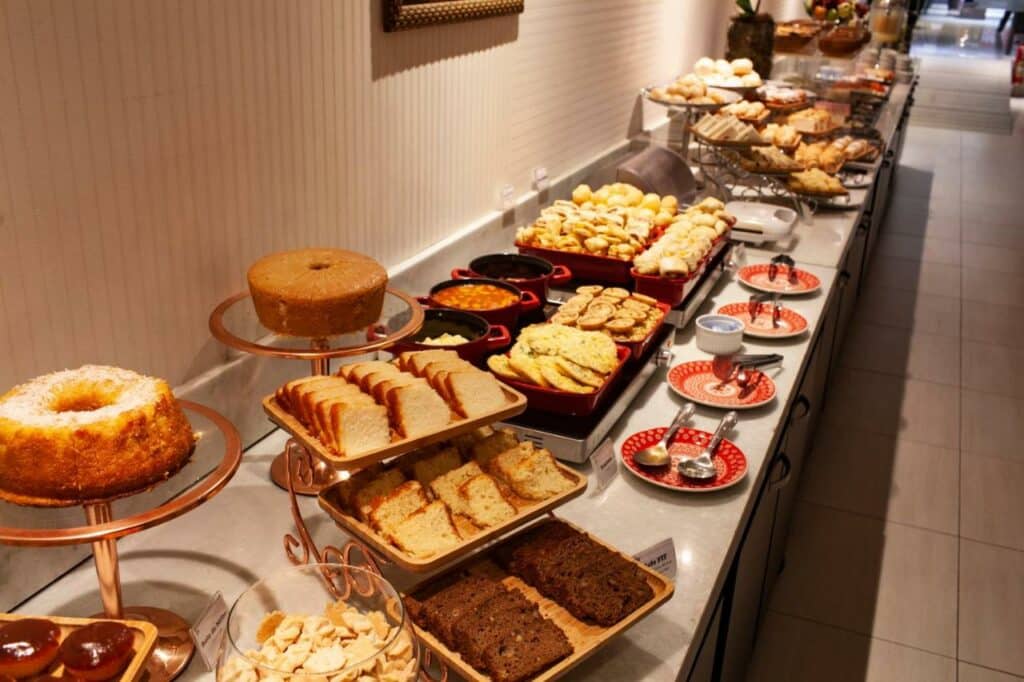 Mesa de café da manhã do Hotel D'Luca com vários tipos de bolos, biscoitos, pães, molhos e pratos.