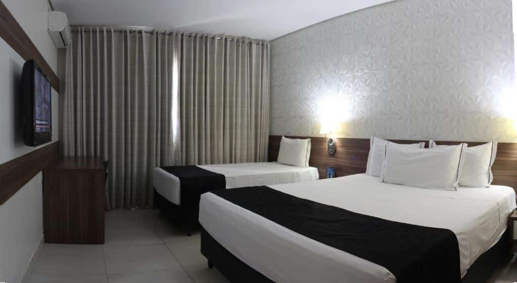 Quarto do Hotel D'Luca com duas camas, uma de casal e outra de solteiro. Há também uma luminária, janela com cortina, mesa e uma televisão. Foto para ilustrar post sobre hotéis em Cuiabá.