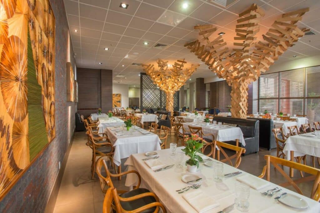 Restaurante do Hotel Gran Odara com várias mesas com cadeiras, vasos de flor, pratos e copos. Ele é todo decorado com material de madeira simulando árvores.