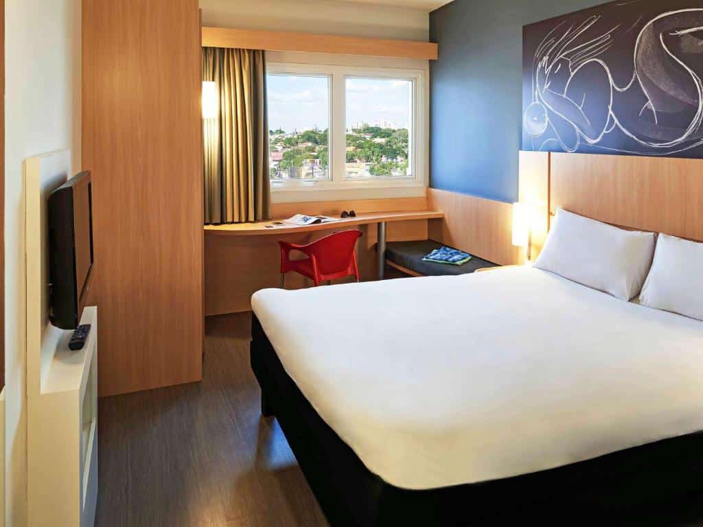 Quarto de hotel Ibis em Goiânia com cama queen-size, móveis de madeira, quadro decorativo em cima da cama, TV em frente a cama, mesa com cadeira em frente a janela e janela com vista para a cidade.