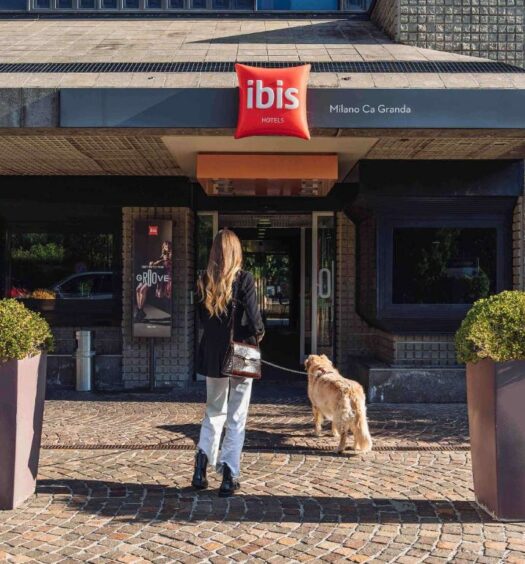 Entrada do Hotel Ibis Milano Ca' Granda, há uma moça de cabelos longos entrando junto o seu cachorro de pelo claro na coleira, para representar hotéis Ibis em Milão