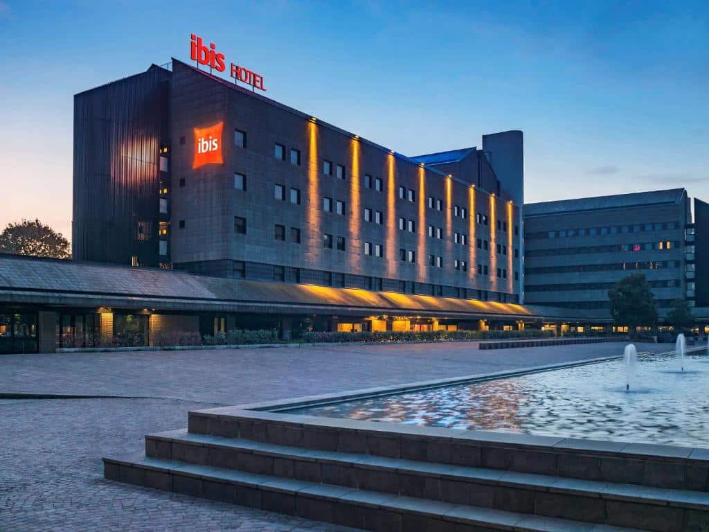 Prédio do Hotel Ibis Milano Ca’ Granda com seis andares em tons de cinza e, na frente do local, há uma ampla fonte, para representar os hotéis Mercure em Milão