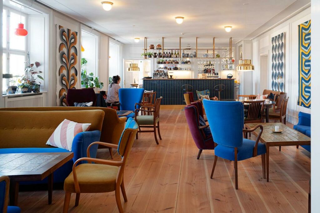 Área comum do hotel Kanalhuset com várias mesas com cadeiras e um bar com várias taças e bebidas. É possível ver também uma pessoa ao fundo sentada em uma dessas mesas.