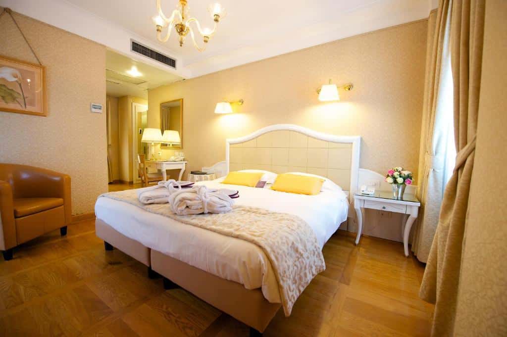 Quarto do Hotel Mercure Milano Centro com uma cama de casal com duas mesinhas de cabeceira com flores, há também uma janela com cortinas, uma mesa de escritório com uma cadeira, uma poltrona acolchoada e um lustre