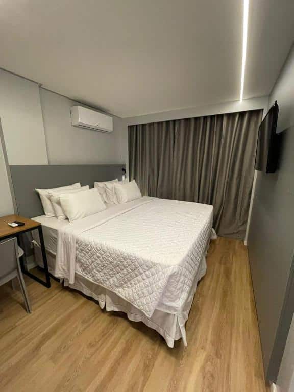 Quarto de hotel minimalista com grande cama de casal com colcha braca, paredes cinza claro e escuro, grande cortina cinza, ar-condicionado e TV. Imagem para ilustrar o post hotéis em Campina Grande.