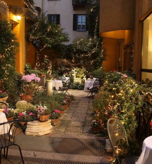 Jardim do Hotel Sanpi Milano, com mesas e cadeiras, vasos de flores com iluminação direta e esculturas, para representar hotéis boutique em Milão
