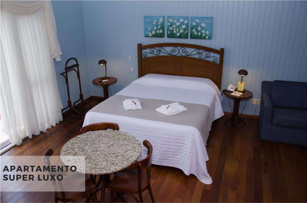 Cama de casal no Hotel Villa Michelon, com cama, quadrinhos sobre a cabeceira, mesinhas com abajures em casa lado, cortina e mesa com cadeiras