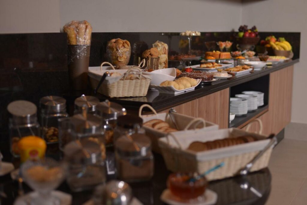 Café da manhã do hotel Hplus Premium Palmas com pães, biscoitos, bolos, frutas e várias outras coisas. Estão todos sobre um balcão que também acomoda alguns pratos.