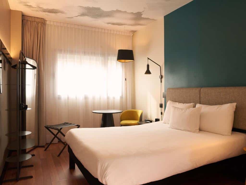 Quarto do Ibis Madrid Aeropuerto Barajas, uma das recomendações de hotéis perto do aeroporto de Madri. Uma cama de casal tem mesinhas de cabeceira e luminárias dos dois lados. Ao fundo fica uma janela com cortinas, e uma mesa com poltrona e abajur está em frente.