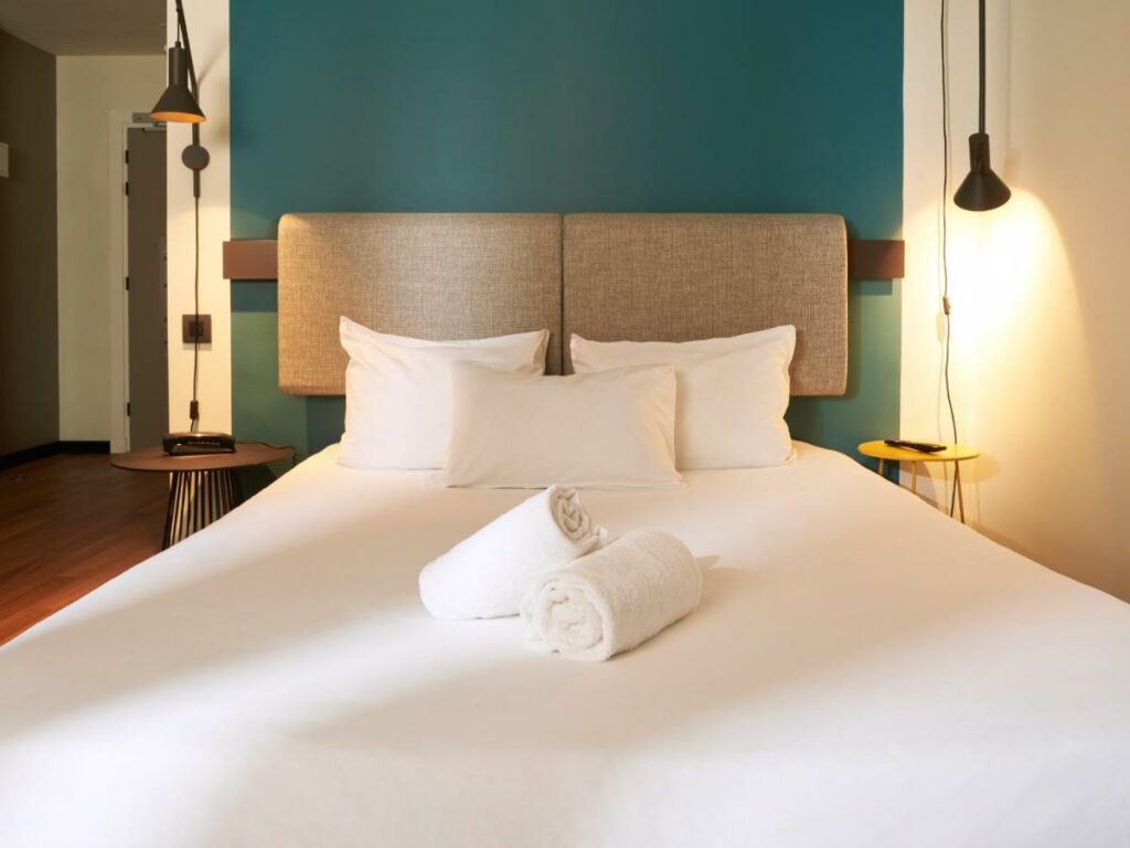 Quarto do Ibis Madrid Aeropuerto Barajas, uma das recomendações de hotéis baratos em Madri. Uma cama de casal tem mesinhas de cabeceira e luminárias dos dois lados. Há duas toalhas brancas enroladas aos pés da cama, e três travesseiros encostados na cabeceira de tecido cinza.