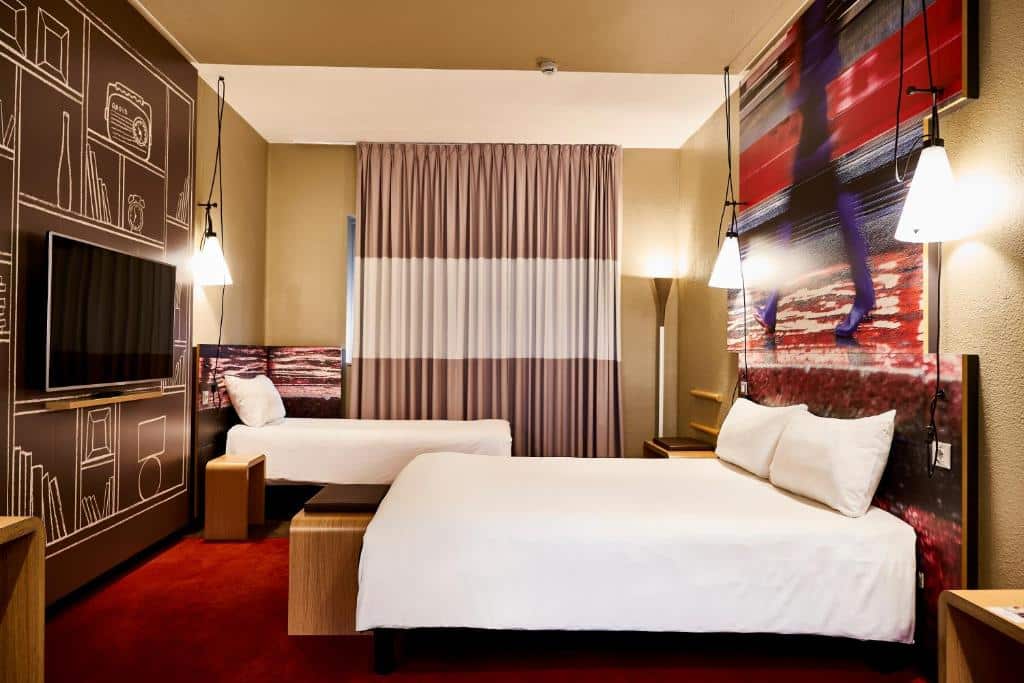 Quarto do Ibis Milano Centro com uma janela ampla com cortinas, há uma cama de casal, uma de solteiro, uma televisão, há também um tapete vermelho e luminárias presas na cabeceira da cama