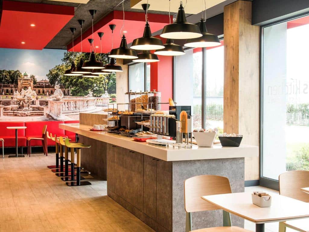 Sala de refeições no Ibis Milano Fiera com um balcão amplo com opções de café da manhã, algumas janelas e sofás vermelhos para sentar-se, para representar hotéis Ibis em Milão