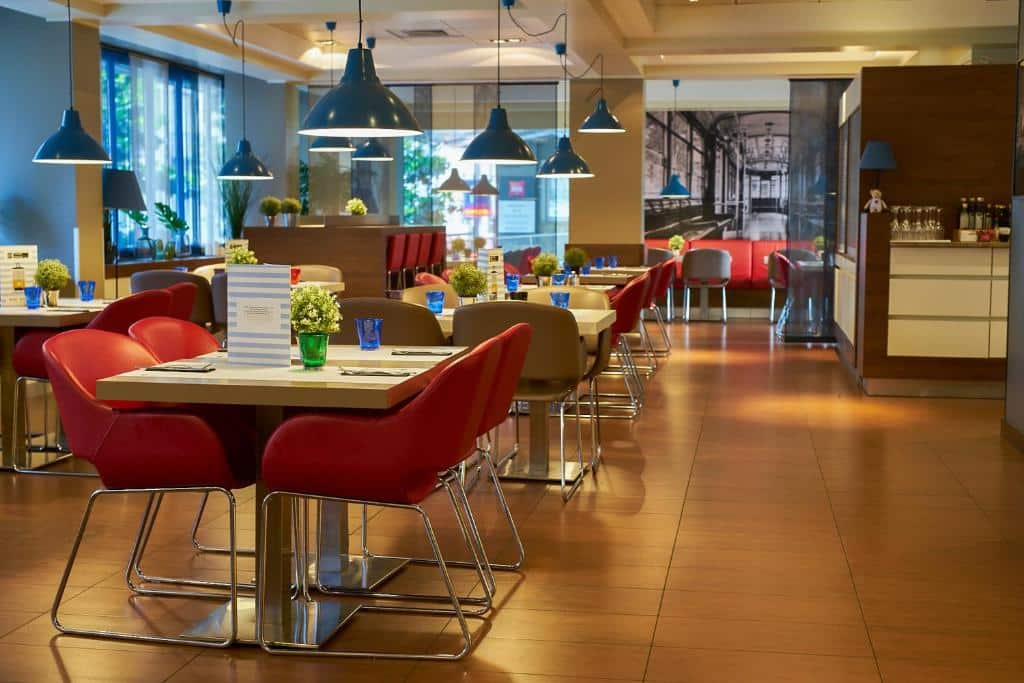 Salão de refeições do Ibis Milano Centro com mesas quadradas, cadeiras vermelhas, luminárias azuis e alguns vasos de plantas também, para representar hotéis Ibis em Milão