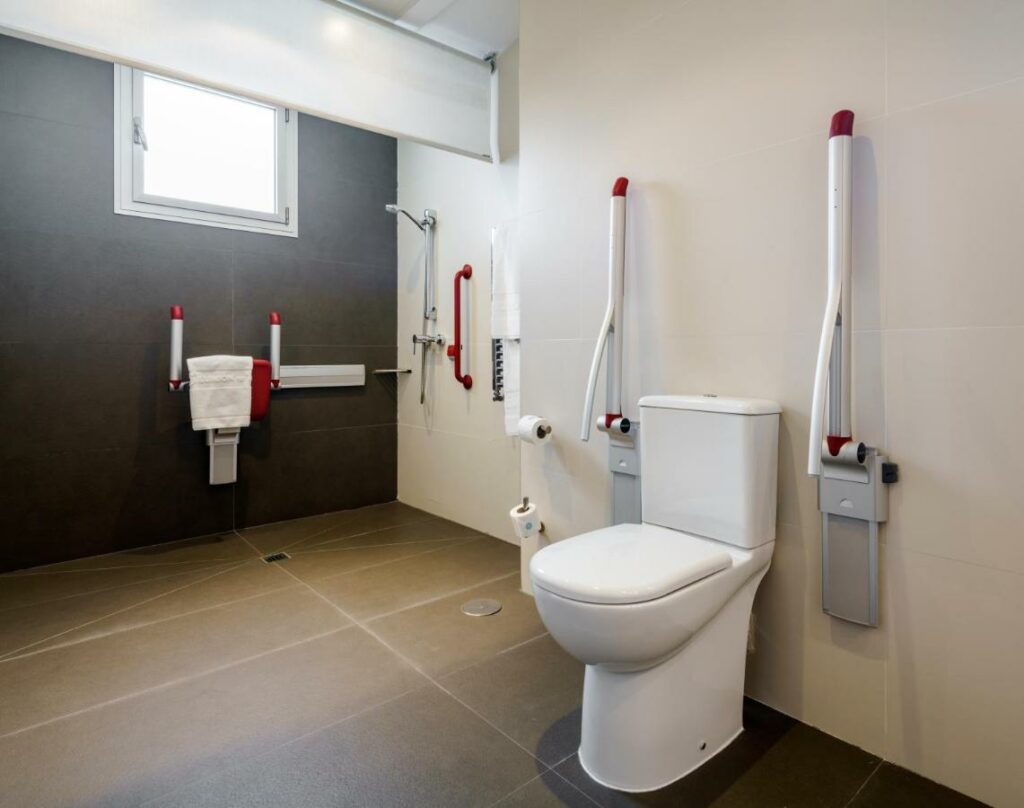 Banheiro adaptado para PCD no Ilunion Alcalá Norte. A privada tem barras de apoio dos dois lados e rolos de papel higiênico entre a área de banho e a privada. Ao lado há barras de apoio ao redor do chuveiro e uma toalha branca pendurada no assento de banho.Há uma janelinha na parede acima.