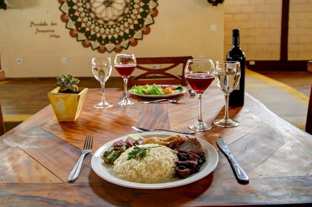Imagem de uma refeição no Hotel Fazenda Colina dos Sonhos, em Socorro. Nos pratos há carne, arroz, salada e outros vegetais. Taças de vinho acompanham a refeição.