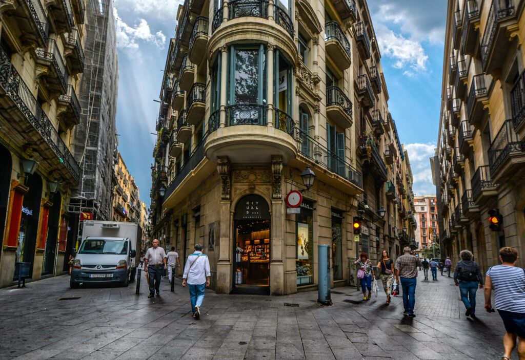 Pessoas caminhando ao redor de prédios no Bairro Gótico para ilustrar o post sobre aluguel de carro em Barcelona. As ruas são bem apertadas, mas há um pequeno caminhão entre os prédios. - Foto: Kirk F via Public Domain Pictures