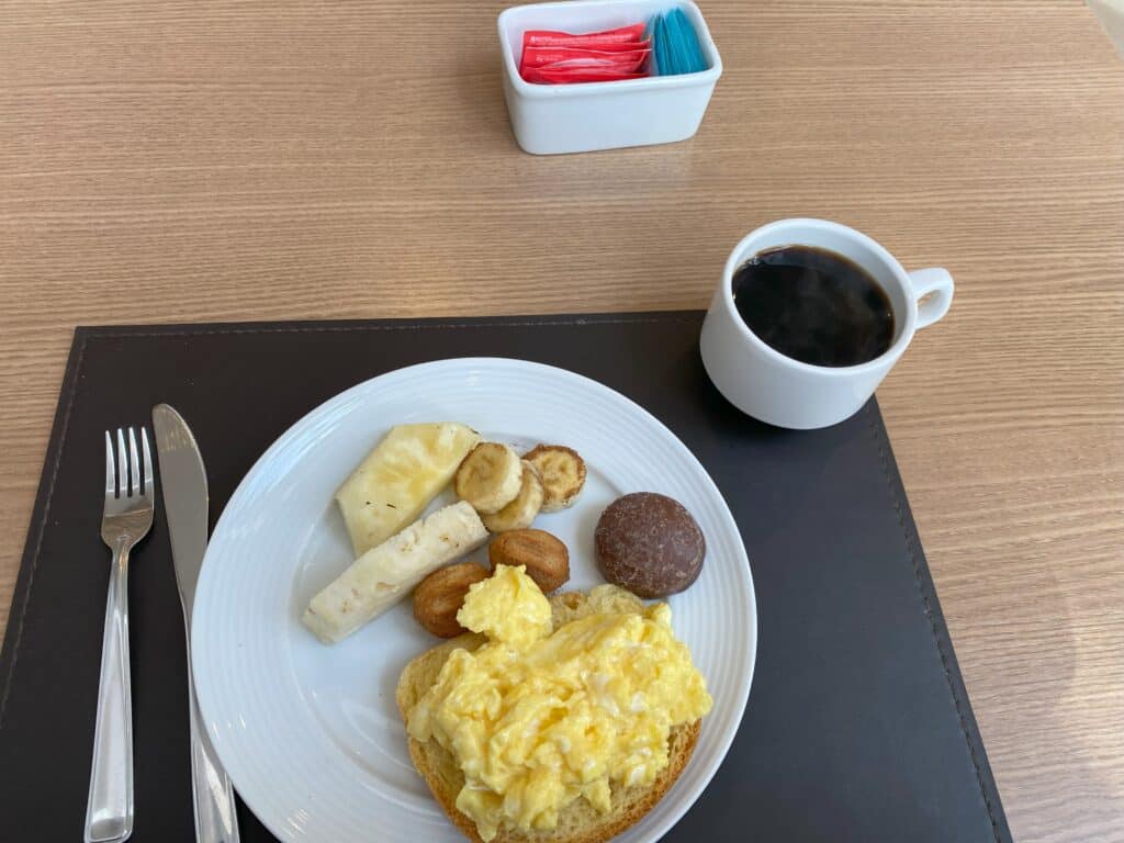 Mesa do café da manhã no Laghetto Viverone Estação, com xícara cheia de café e prato servido com pão, ovos mexidos, dois mini churros, um pão de mel, duas metades de uma fatia de abacaxi e três rodelas de banana com canela