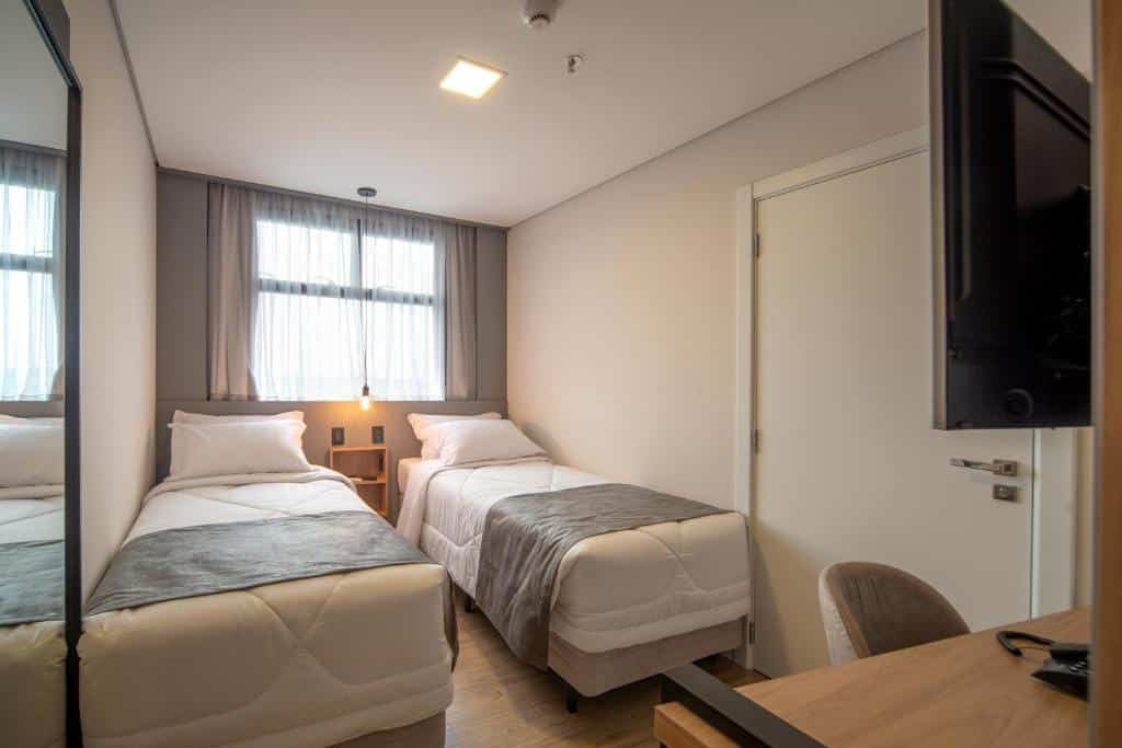 Duas camas de solteiro, porta, cortina na janela, pendente de luz e TV sobre a mesa de trabalho com cadeira no quarto Standard do hotel