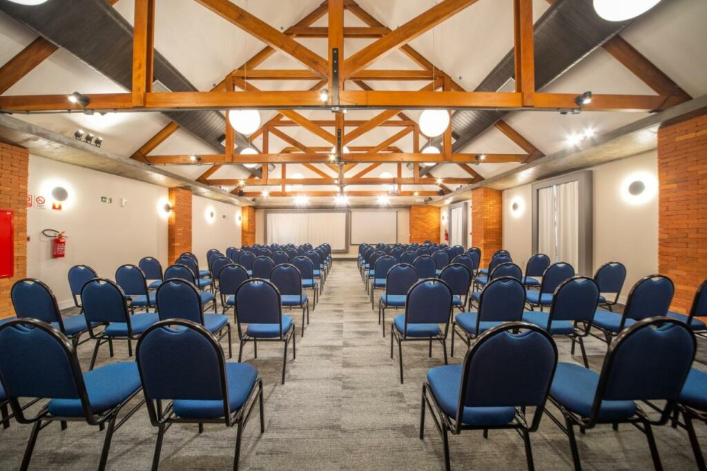 Sala de eventos do hotel, com detalhes em madeira no teto, paredes com colunas de tijolos à vista, além de cadeiras com estofado azul organizadas de modo a deixar um corredor entre elas