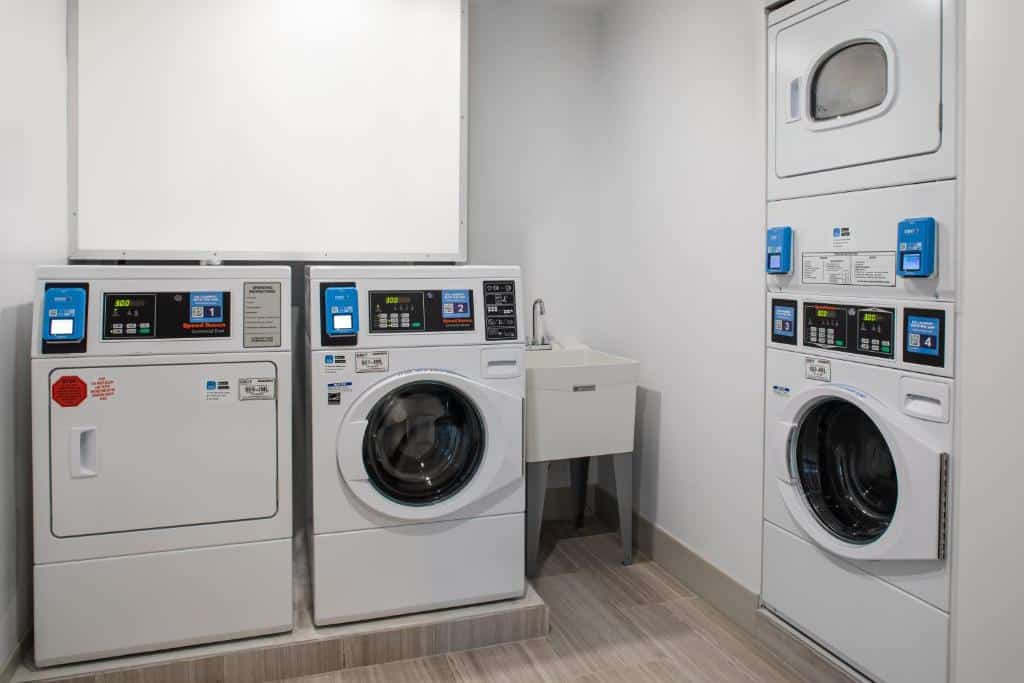 lavandeira do Holiday Inn Express & Suites com duas máquinas de var e duas secadoras, todas brancas. Duas delas estão no chão, as outras duas estão uma em cima da outra na parede, no lado direito