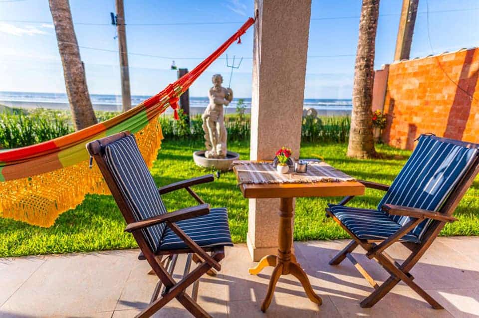 No Mar Egeu Arte & Pouso. Uma mesa com duas cadeiras de madeira no lado direito. Atrás uma rede com uma área verde e vista para a praia.