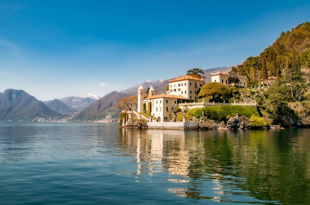 Lago Como, um extenso lago cercado por montanhas e muito verde, em destaque na imagem, há uma construção pintada de branco às margens do lago