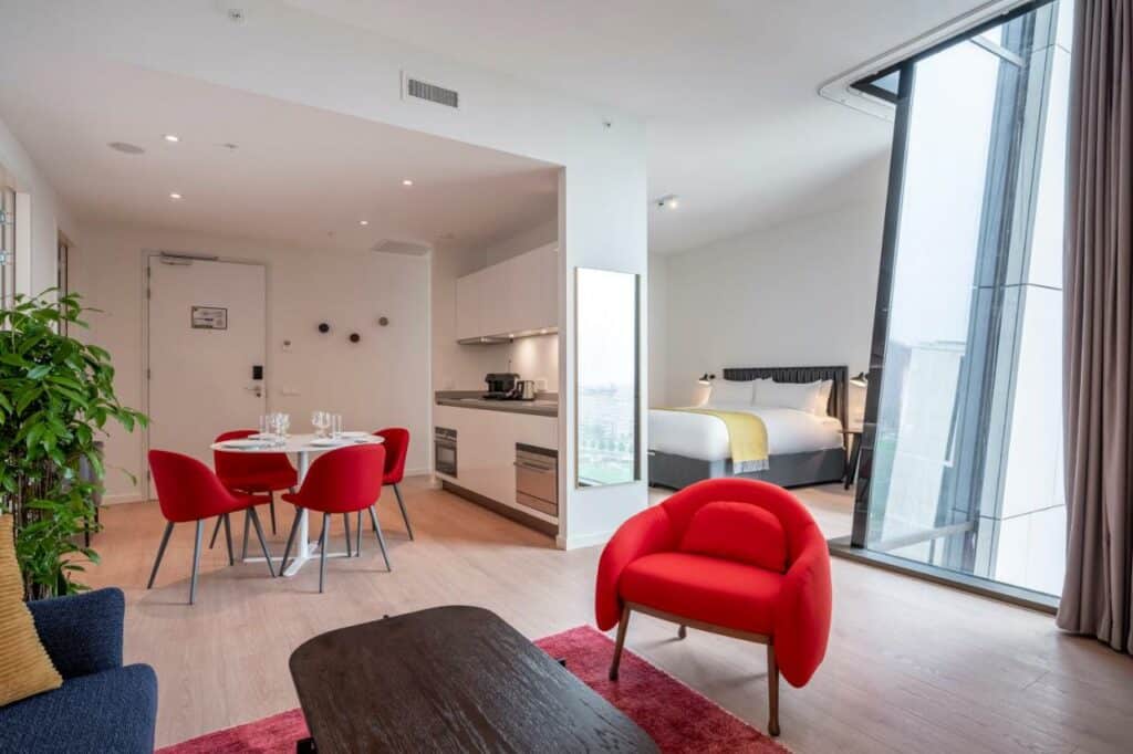 Loft do Premier Suítes Plus Amsterdam, contendo uma poltrona vermelha, uma mesa de centro de madeira e um tapete vermelho em baixo. Ao fundo tem a área da cozinha à esquerda, com uma mesa redonda e cadeiras vermelhas e a pia. À direita, há uma cama de casal