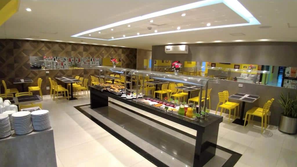 Parte do hotel para fazer as refeições, ao centro um balcão com as comidas e em volta mesas com cadeiras amarelas, ilustrando post Hotéis em Maringá.