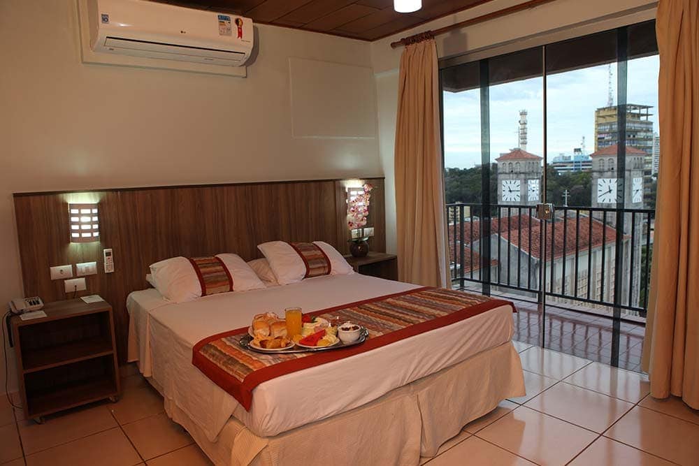 Quarto do Mato Grosso Palace Hotel com uma cama de casal com café da manhã em cima, ar-condicionado e uma varanda com vista da cidade. Foto para ilustrar post sobre hotéis em Cuiabá.