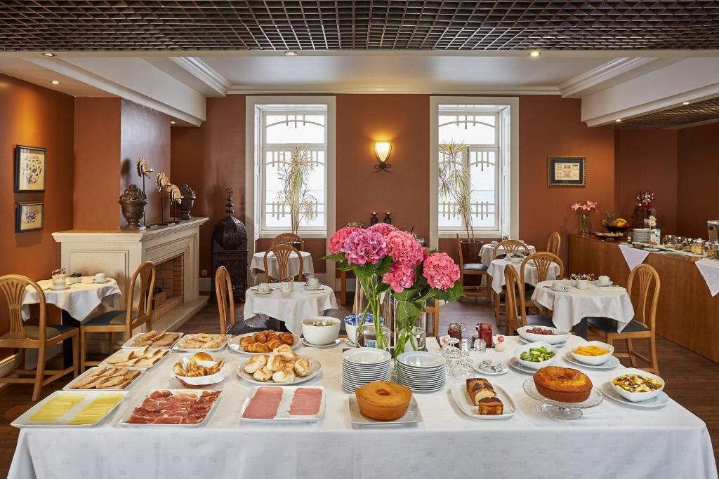 Área de refeições em hotel com arquitetura clássica, mesa de café da manhã com toalha branca, pratos com bolos, frios, pães, mesas com cadeiras ao fundo e duas janelas.