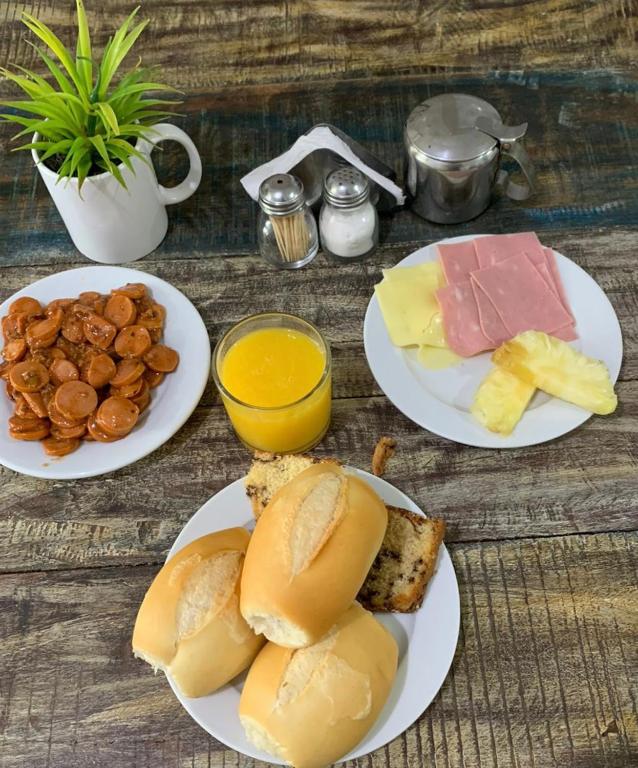 Mesa de madeira com pratos com café da manhã com pães, bolos, salsicha, queijo, presunto, abacaxi, copo com suco e pequeno vaso com planta decorativa.