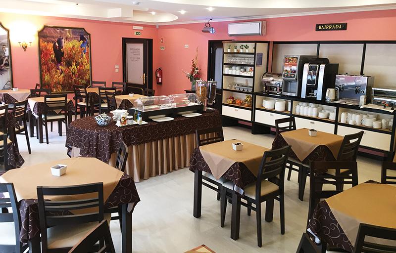 Área de refeições em hotel, com paredes rosa, mesas e cadeiras marrons, máquinas de café em uma estante e mesa para servir alimentos no meio da sala.