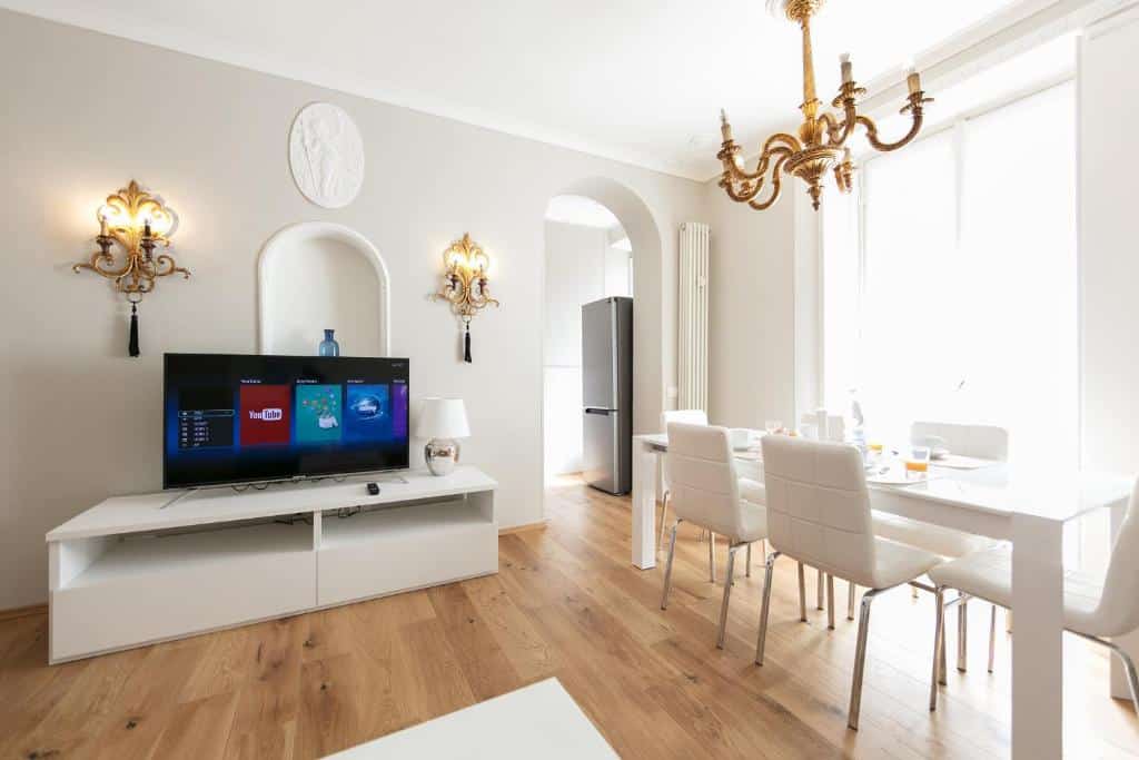 Sala de estar do  Milan Retreats com um rack com televisão, uma mesa com seis lugares, um lustre dourado, piso que imita madeira e as paredes são brancas