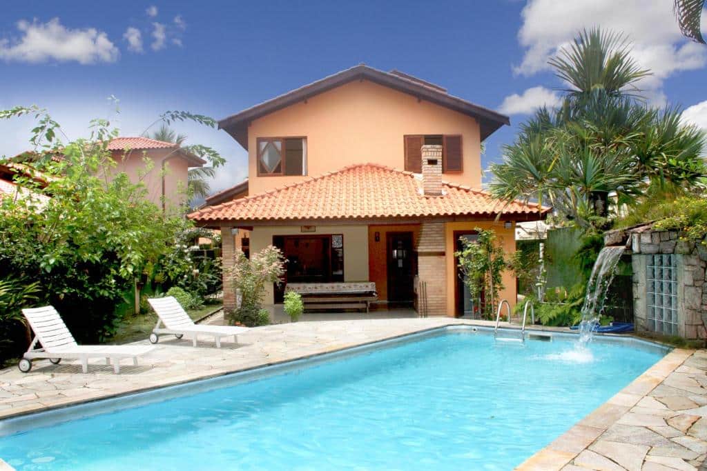 Casa Morada Canavieiras. Uma piscina na frente, atrás duas cadeiras de tomar sol e o fundo da casa com churrasqueira e árvores.