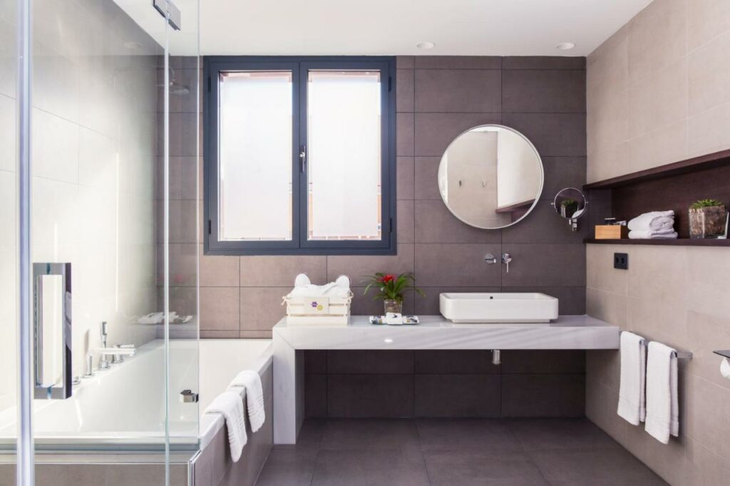 Banheiro do Motel One Madrid-Plaza de España. Uma banheira está em frente ao box com chuveiro e ao lado da bancada com toalhas e pia. Há um espelho redondo e uma janela na parede acima da bancada.