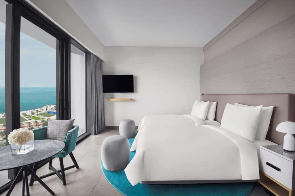 Quarto do Mövenpick Resort Al Marjan Island com duas camas de solteiro, uma varanda ampla com vista pro mar, uma televisão e uma pequena mesa redonda com uma cadeira estofada
