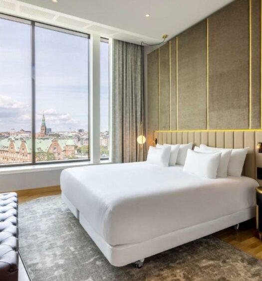 Quarto do hotel NH Collection Copenhagen com uma cama de casal, mesinha de cabiceira, televisão, um divá encostado na parede e uma janela grande com vista para a cidade. Foto para ilustrar o post hotéis em Copenhague.