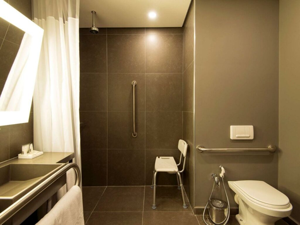 Banheiro acessível do Novotel Santos Gonzaga. Há barra de apoio acima da privada (que é mais baixa), na área do chuveiro e na pia. Há também uma cadeira de banho.
