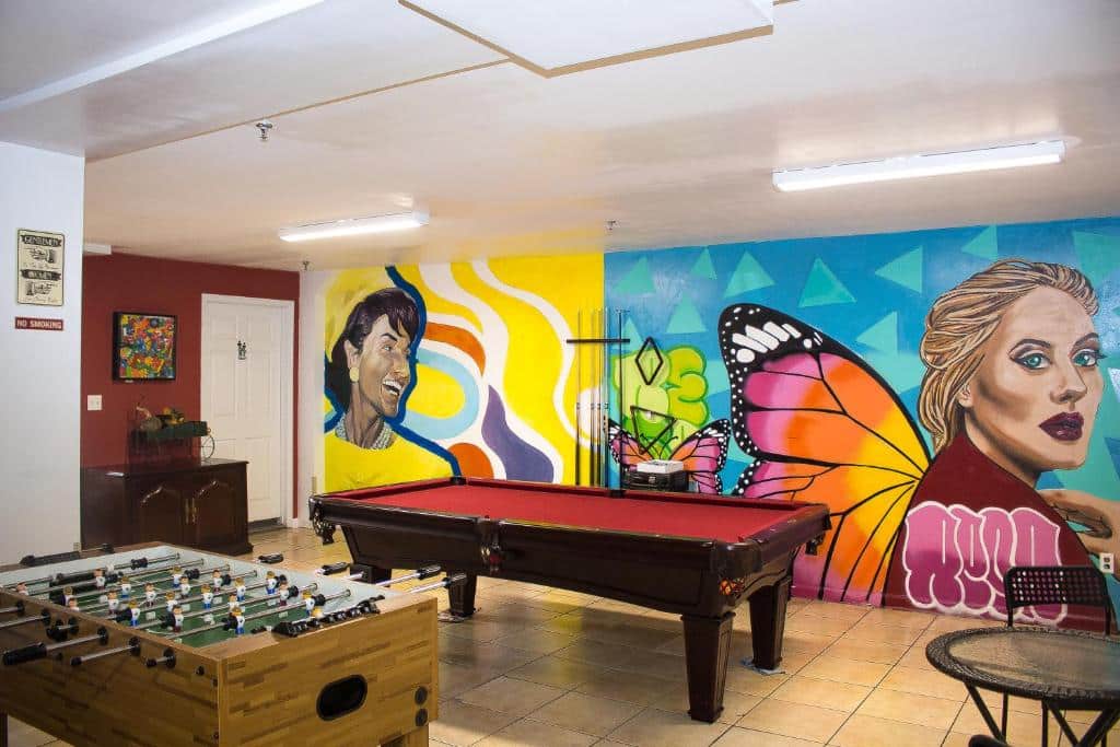 área de recreação do NY Moore Hostel com uma mesa de pebolim, uma mesa de sinuca e uma parede de fundo toda pintada e grafitada com cores chamativas, dentre as pessoas pintadas, está a imagem da cantora Adele.
