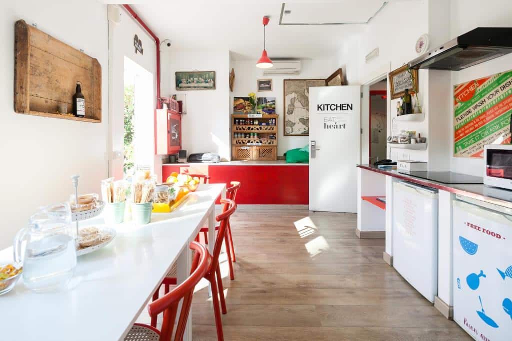 Cozinha compartilhada do Ostello Bello cm uma ampla mesa com cadeiras vermelhas, há balcões com itens de café da manhã, além de um frigobar e um microondas