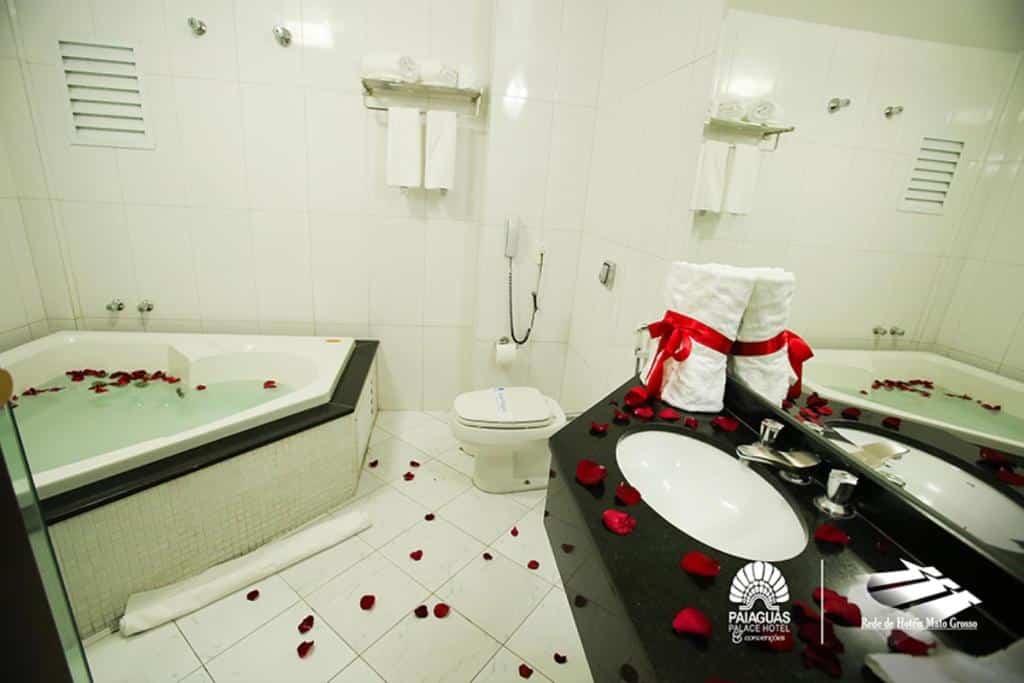 Banheiro do Paiaguas Palace Hotel com uma banheira de hidromassagem em formato de coração, pia, vaso sanitário e toalhas. Pétalas de rosa estão espalhadas por todo o banheiro.