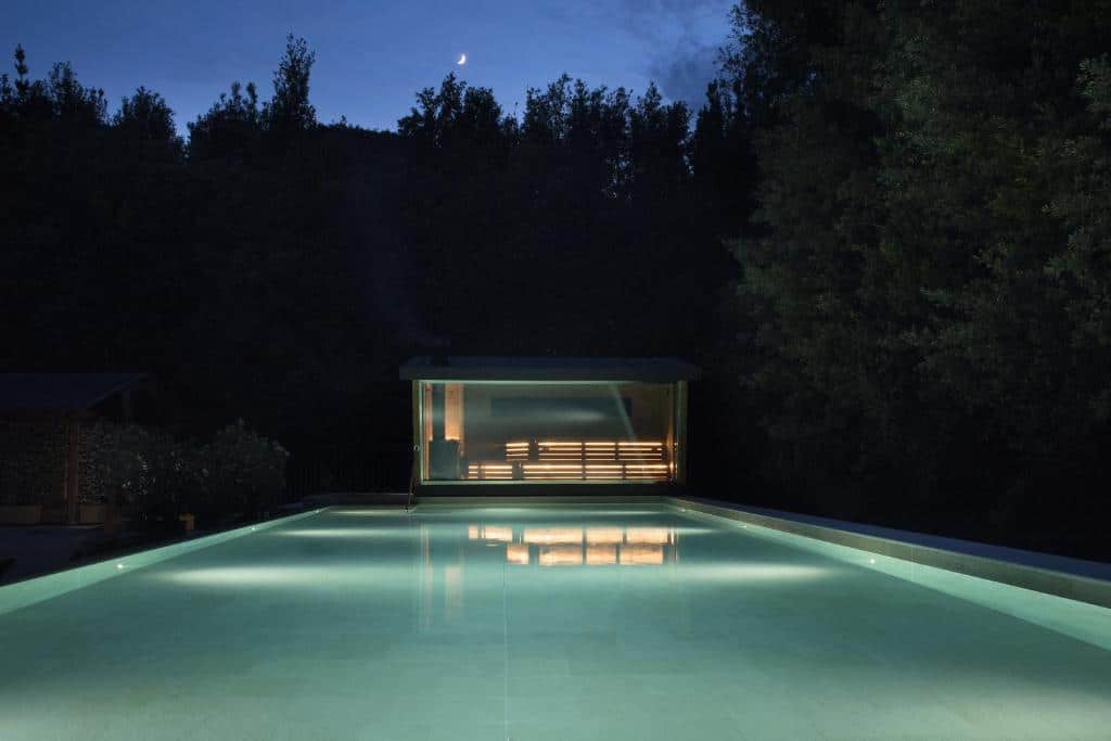 piscina do QC Termeroma Spa and Resort à noite bem iluminada, cercada de natureza e, ao fim dela, a sauna com paredes de vidro e iluminada
